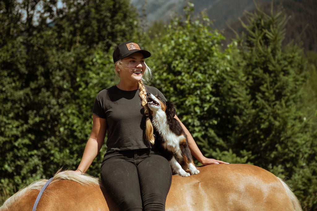 Woman sitting on a horse with a dog on her lap, surrounded by greenery
